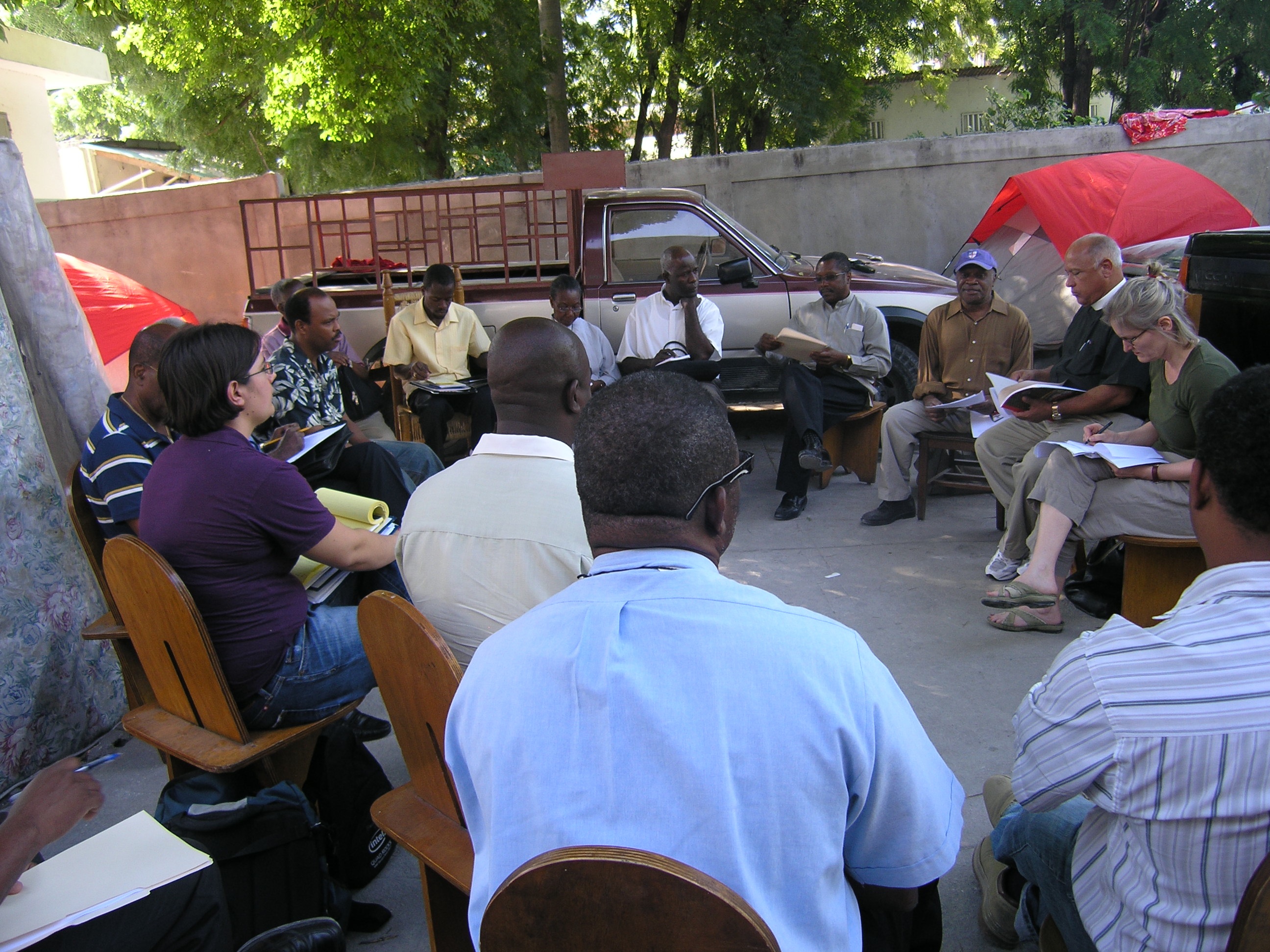 Group of women and men in a circle of wooden chairs in and outdoor lot